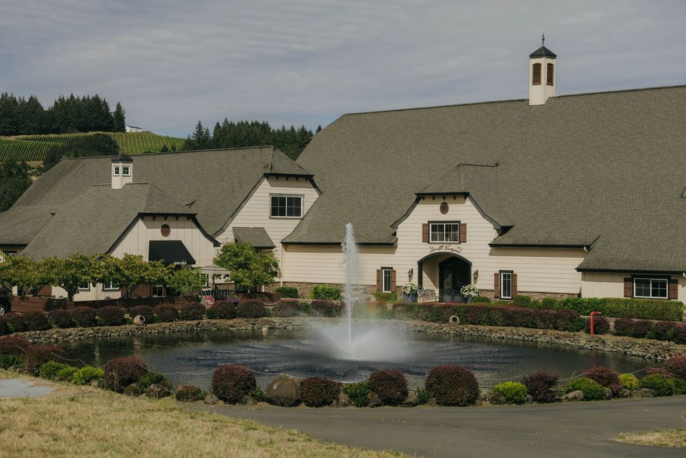 Elegant winery estate with fountain pond, manicured gardens, and white stucco building with gray roof in wine country.