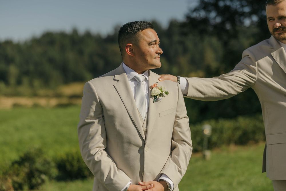 Groom in beige suit with floral boutonniere being congratulated outdoors at a sunny wedding ceremony.