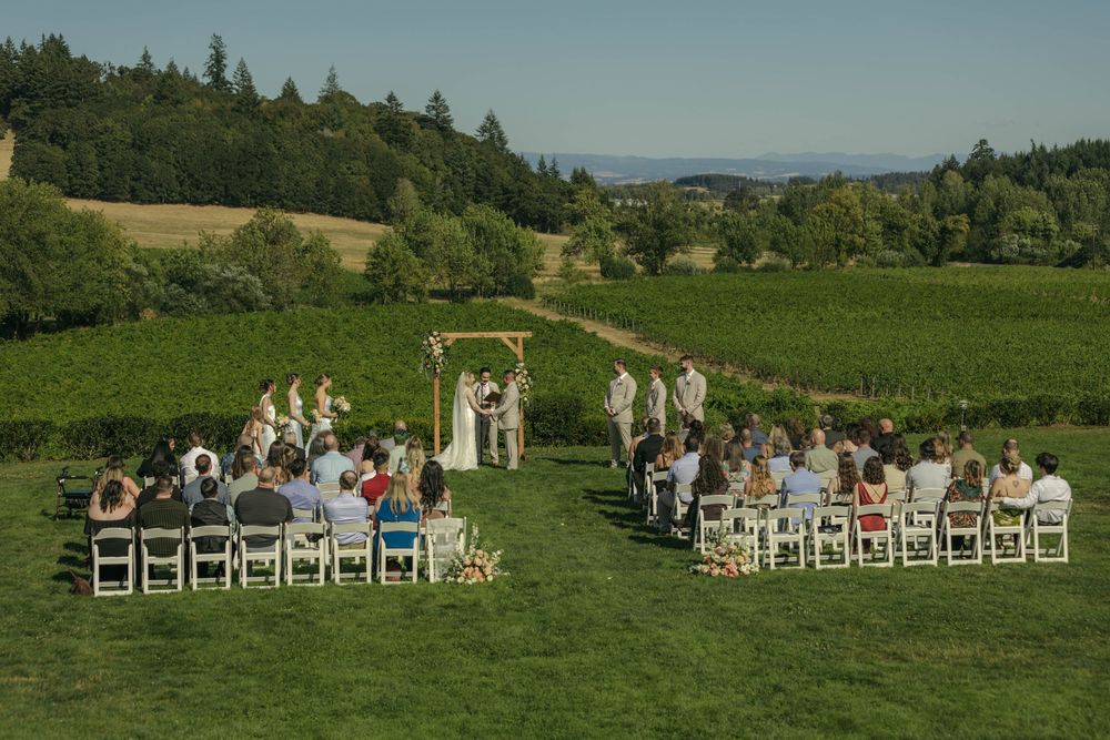 Outdoor wedding ceremony in a lush green vineyard with guests seated in white chairs beneath a wooden arch.