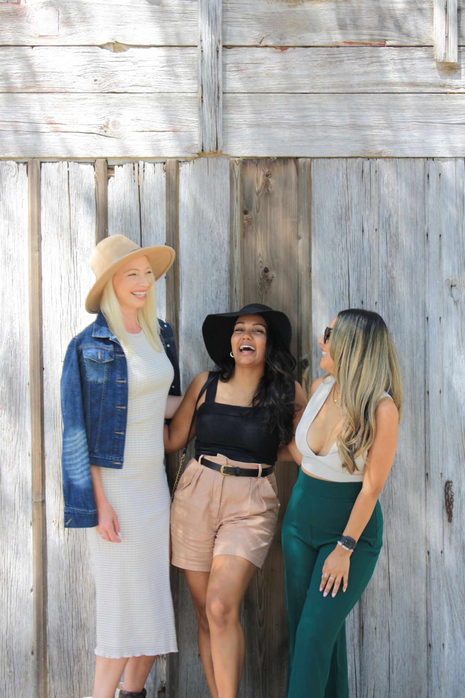 Three friends in stylish outfits sharing laughs against rustic wooden wall backdrop.