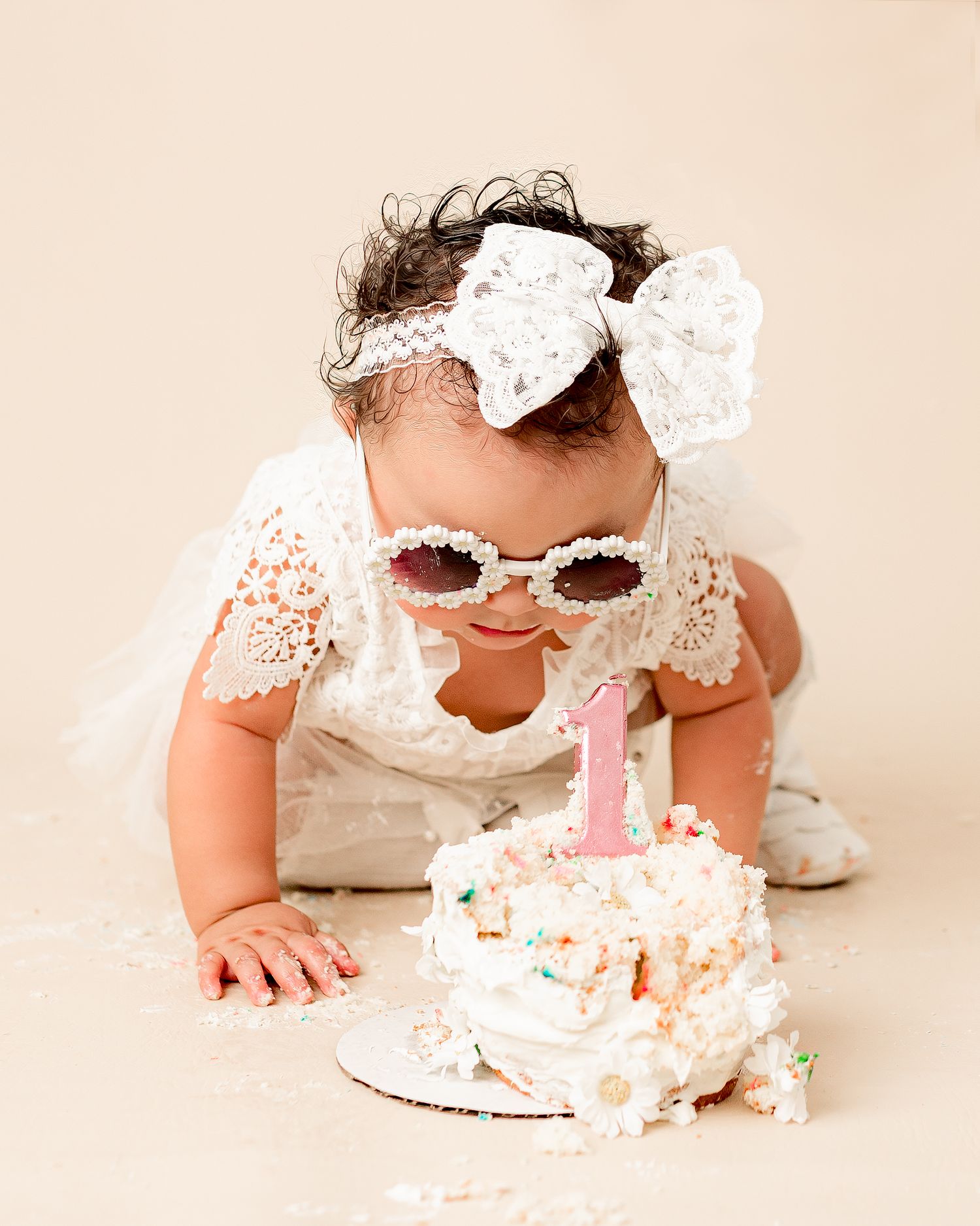 Baby wearing white lace gloves and sunglasses plays with frosted birthday cake during first birthday smash cake session.