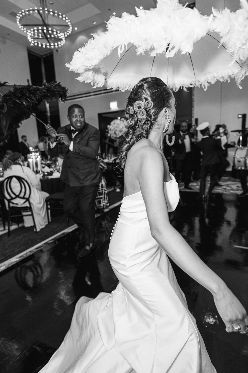 A festive wedding dance celebration under decorative white feathered umbrellas and crystal chandeliers.