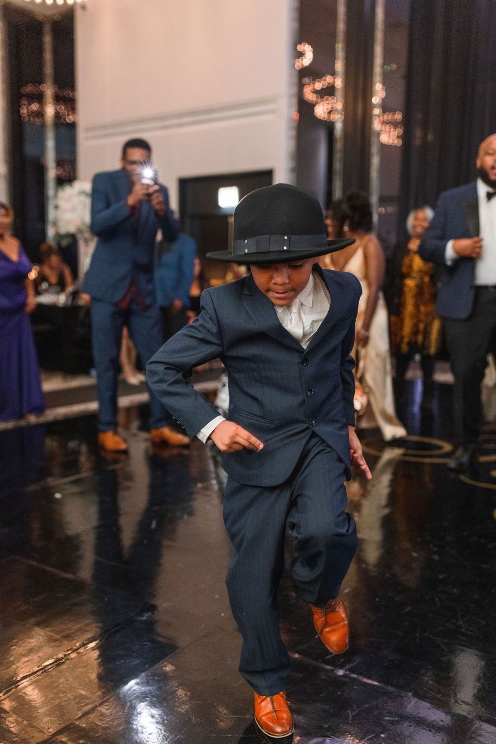 Young guest in suit and hat performs solo dance at wedding reception.