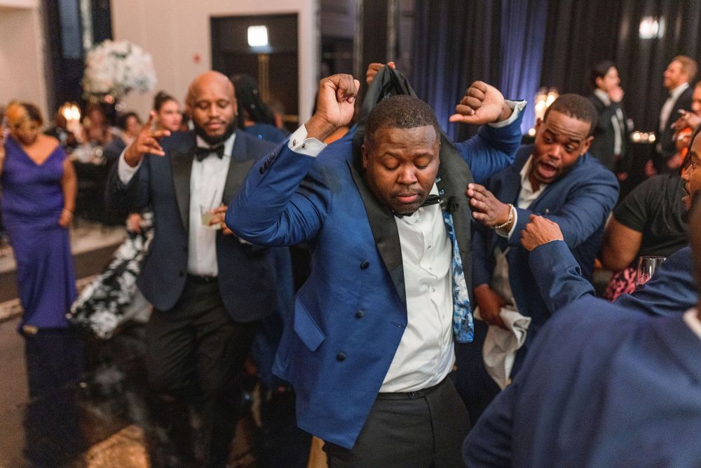 Wedding guests dance enthusiastically in blue suits during a lively reception celebration.