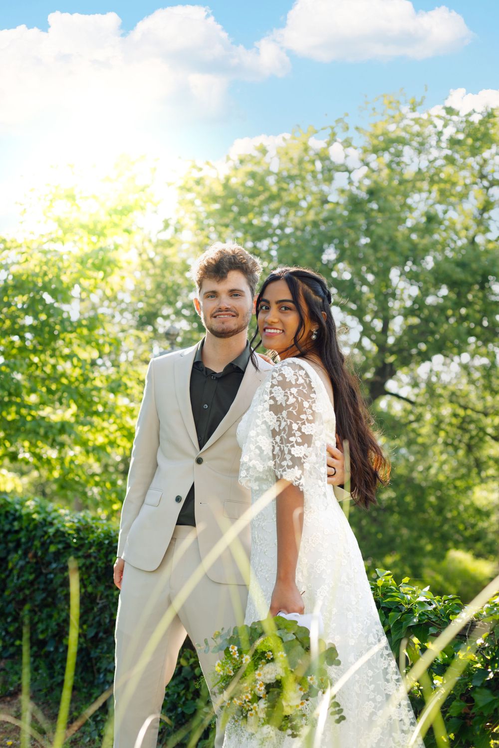 Romantic wedding couple poses together outdoors with lush greenery and sunny blue sky in background.