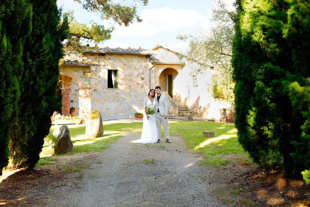 Newlyweds walk hand in hand along a rustic stone path surrounded by cypress trees.