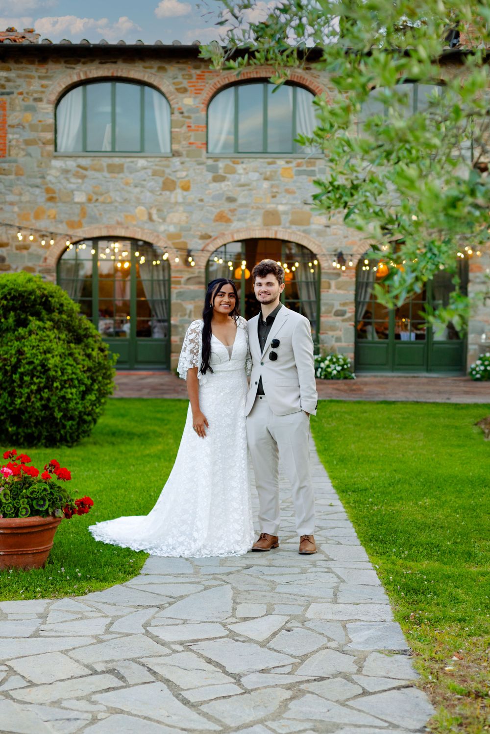 Elegant couple in white attire poses on stone pathway in front of rustic Tuscan villa.