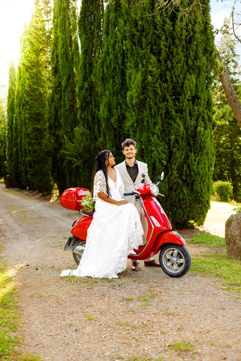 Wedding couple posing with red Vespa scooter alongside row of cypress trees.