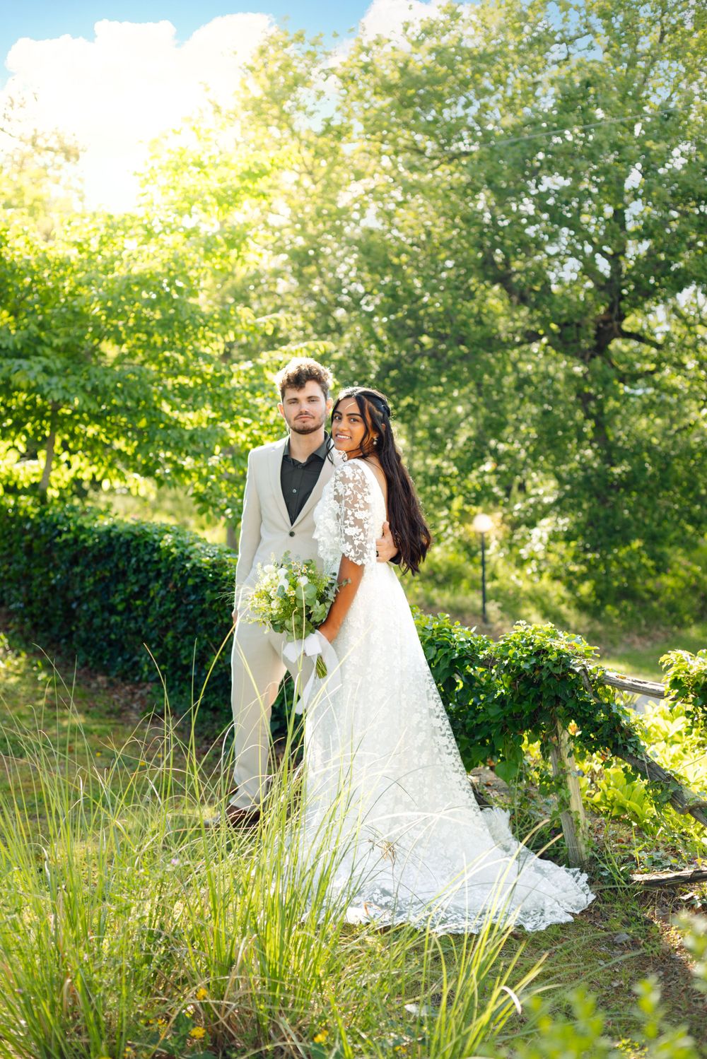 Wedding couple standing in sunlit garden with lush greenery backdrop.