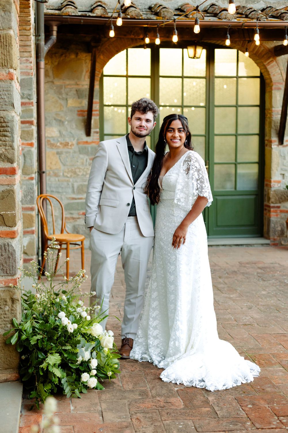 Wedding couple poses beneath string lights outside rustic stone building with elegant windows.