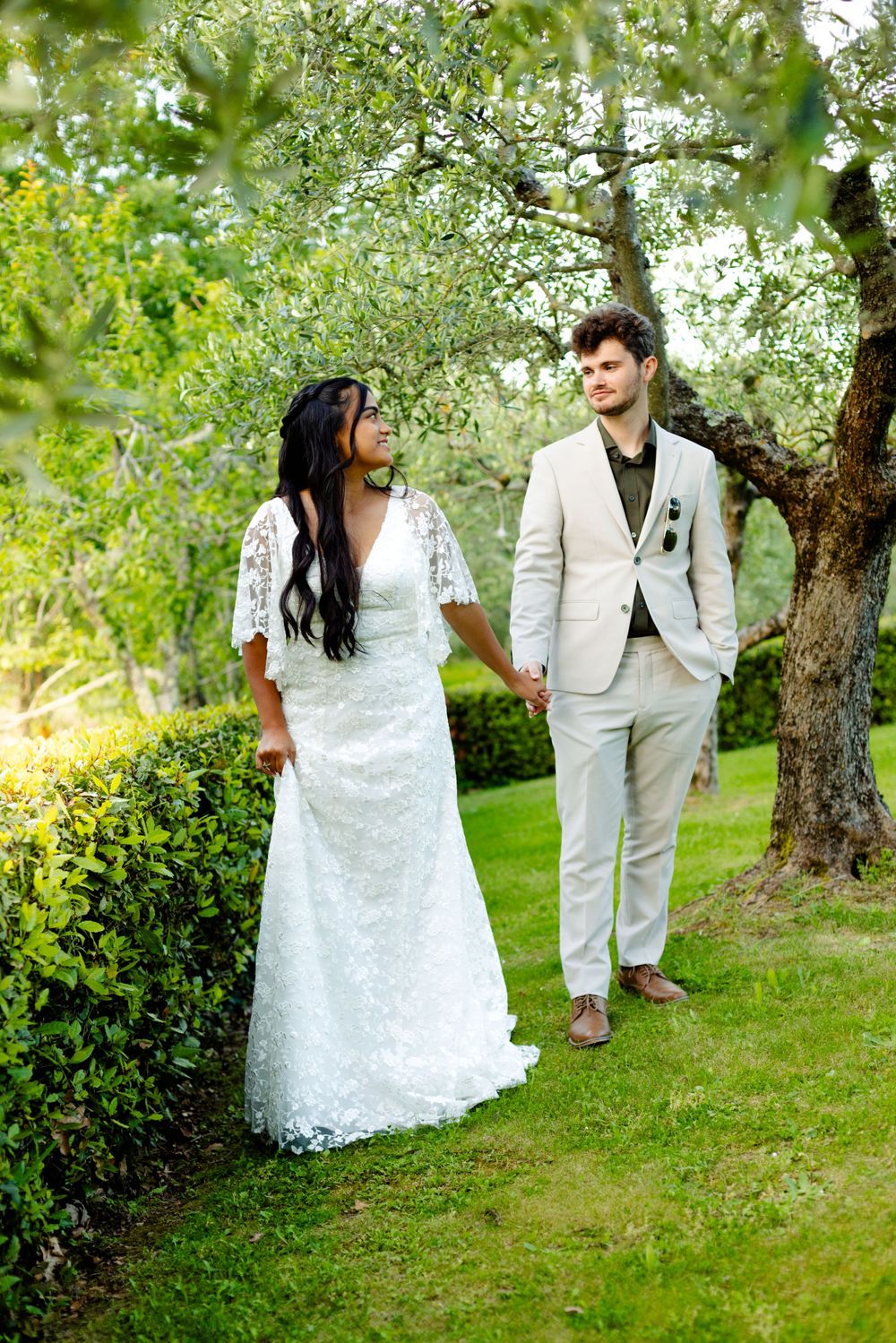 Couple in white wedding attire walks through scenic garden with blooming trees.