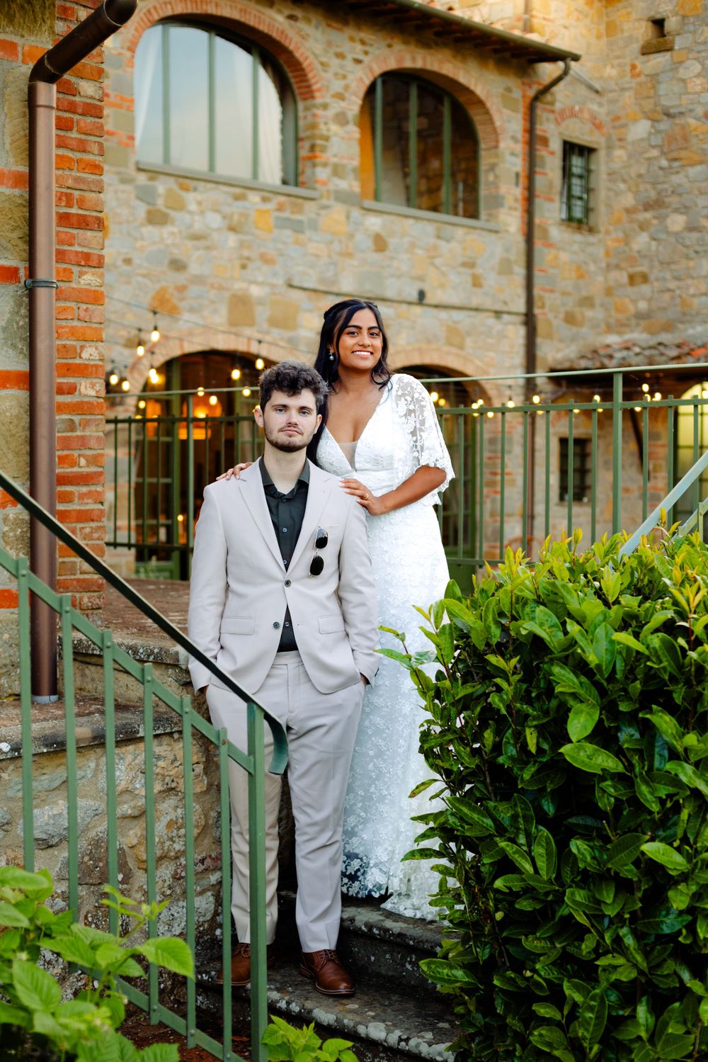 A couple poses on stone steps outside a rustic Tuscan villa with arched windows and string lights at dusk.