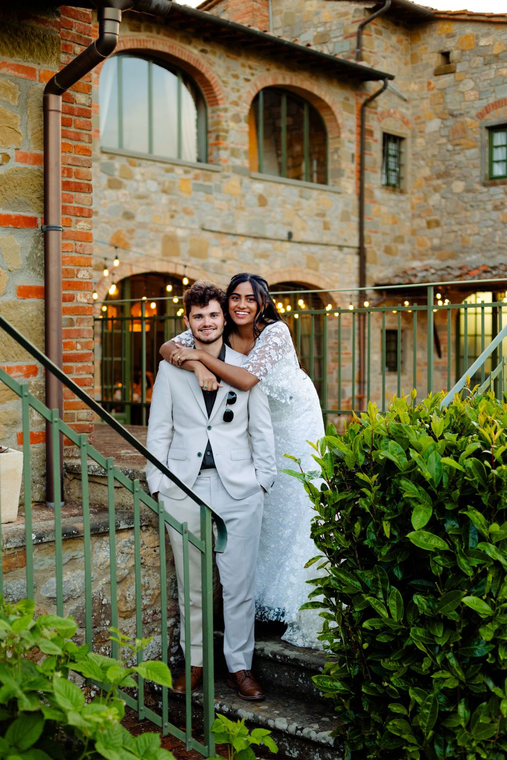 A happy couple embraces on rustic stone steps surrounded by greenery and string lights.