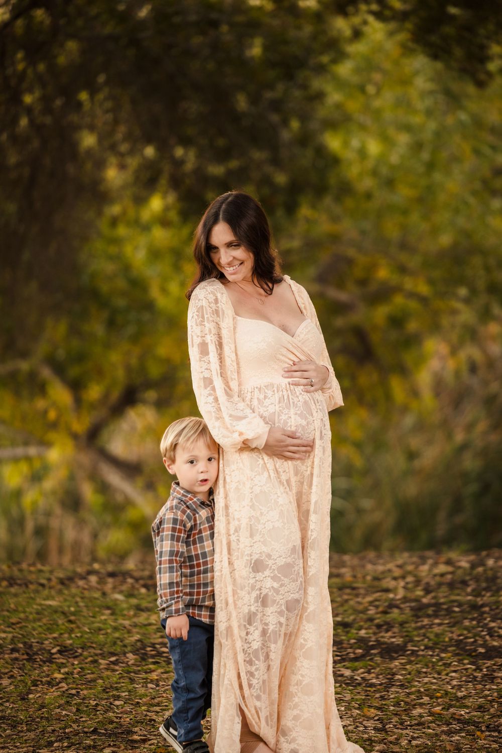 A maternity photo outdoors showing a person in a cream lace dress with a young child in plaid and jeans.