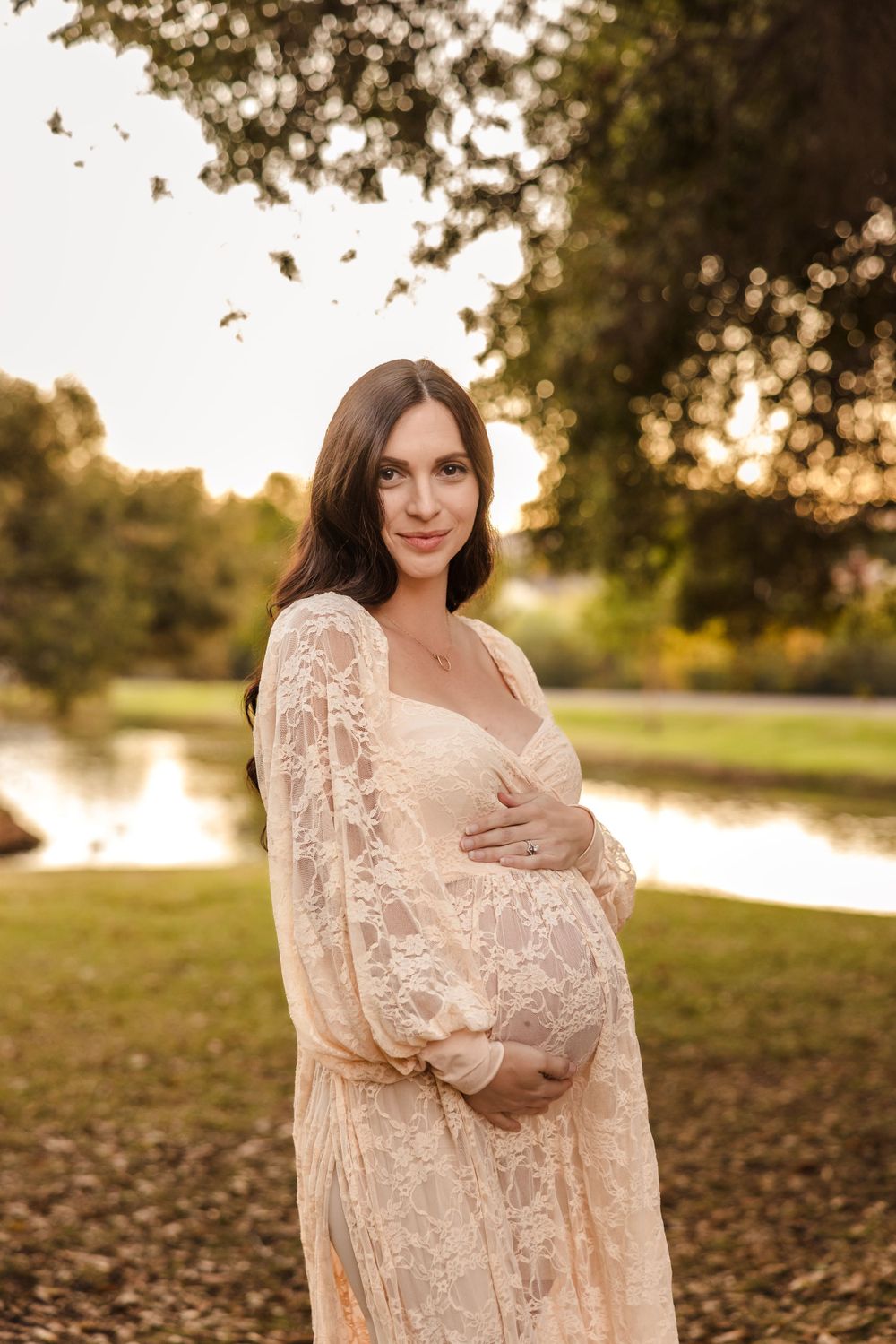 A maternity portrait by a lake at sunset featuring a cream lace dress and natural setting.