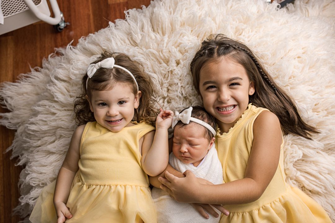 Three siblings lying on a fluffy white rug, wearing yellow dresses and white outfit for a sweet portrait session.