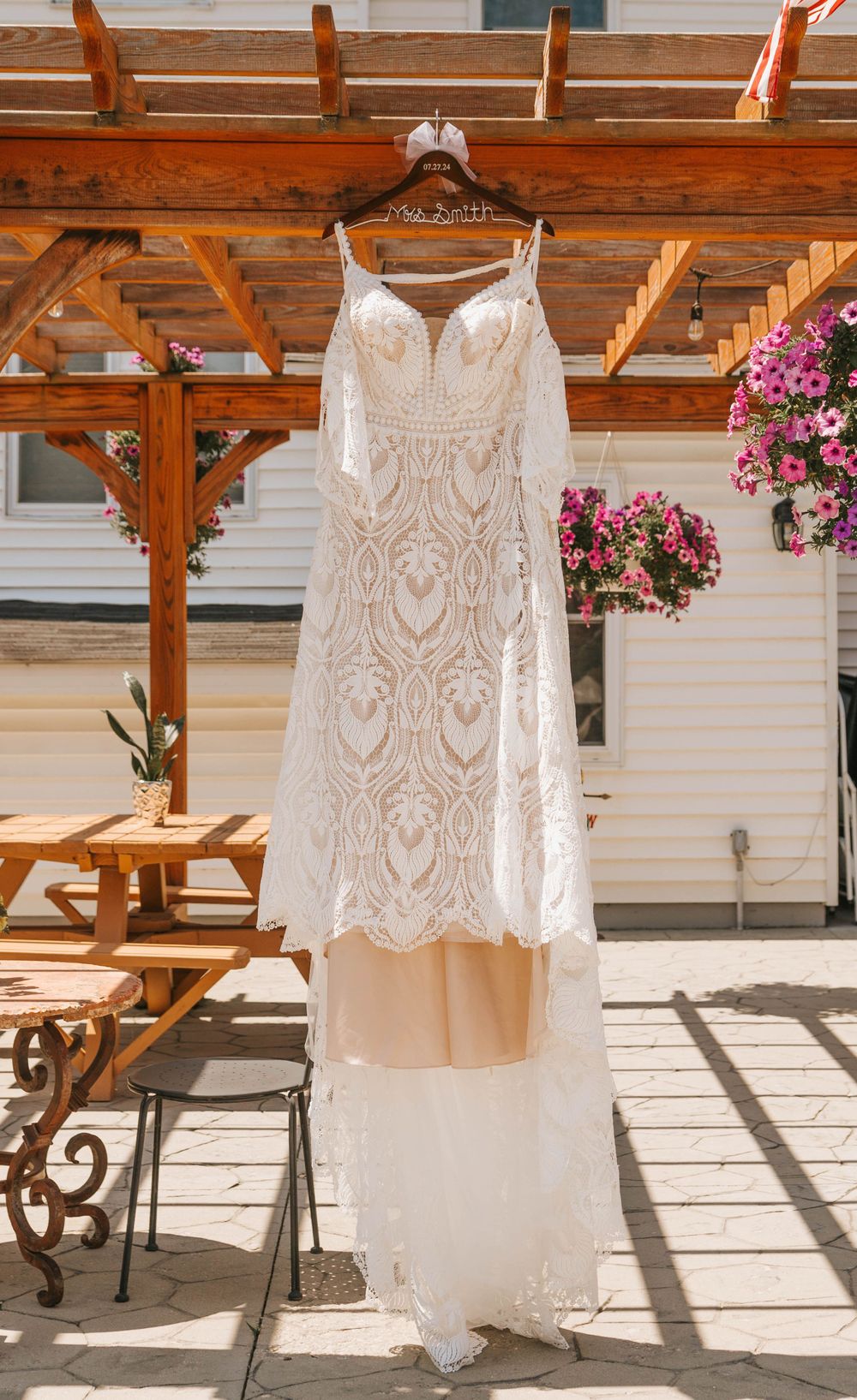 Elegant white lace wedding dress hanging on wooden pergola with pink flowers in background.