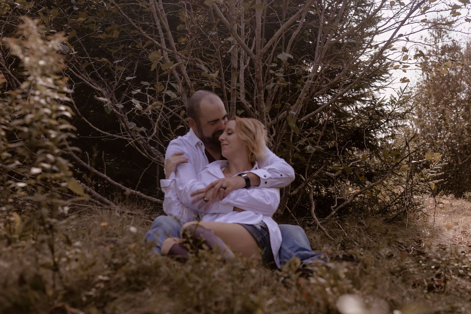 Romantic couple in white shirts embracing on grassy hillside among trees.