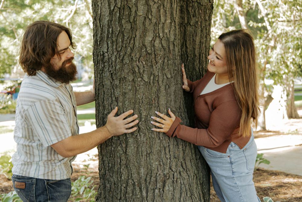 A couple hugs a large tree trunk together in a playful outdoor moment.