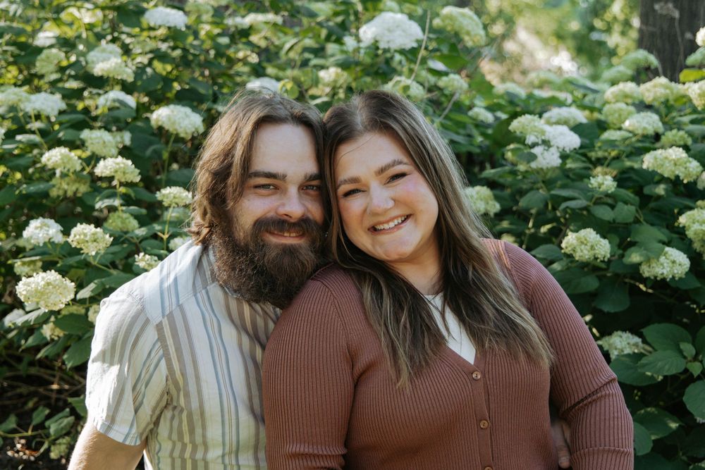 A couple poses together in front of blooming hydrangea bushes on a sunny day.