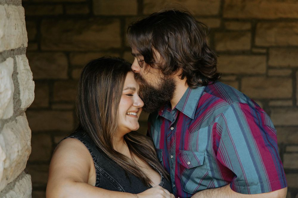 Couple sharing an intimate moment against a rustic stone wall background in casual attire.