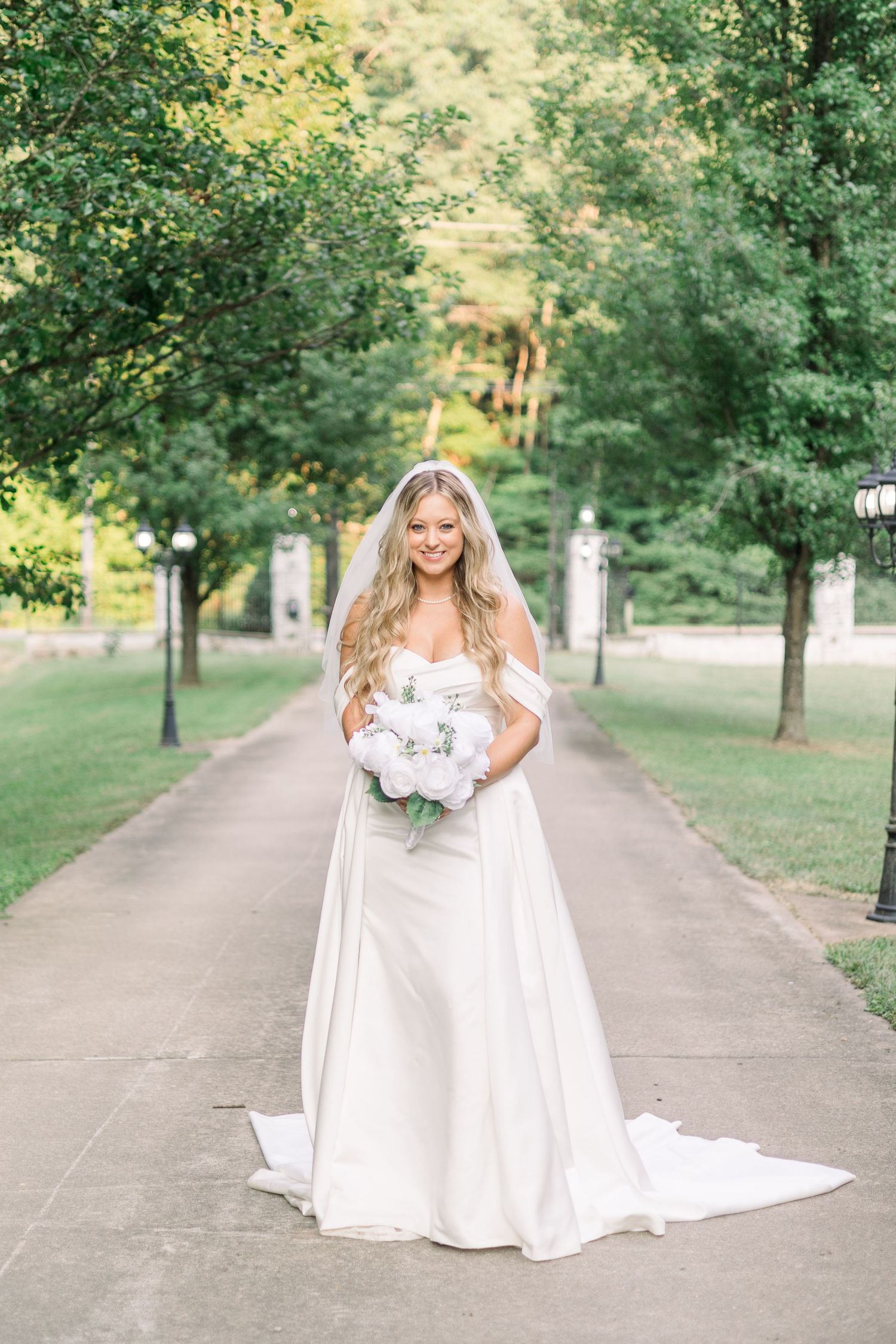 Bride in flowing white gown poses on tree-lined pathway during outdoor photoshoot.
