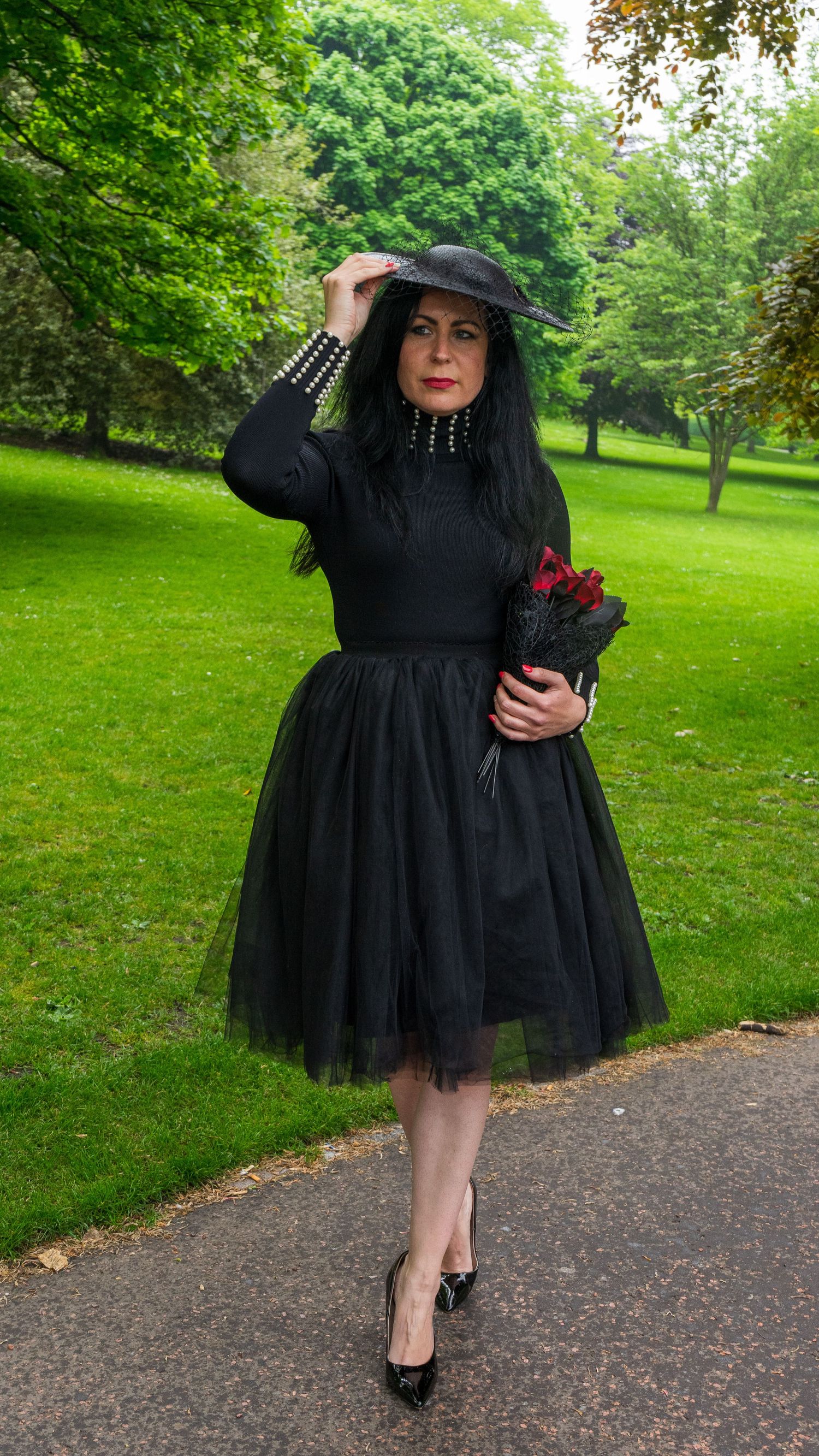 A model in a black dress poses on a park pathway surrounded by green grass.
