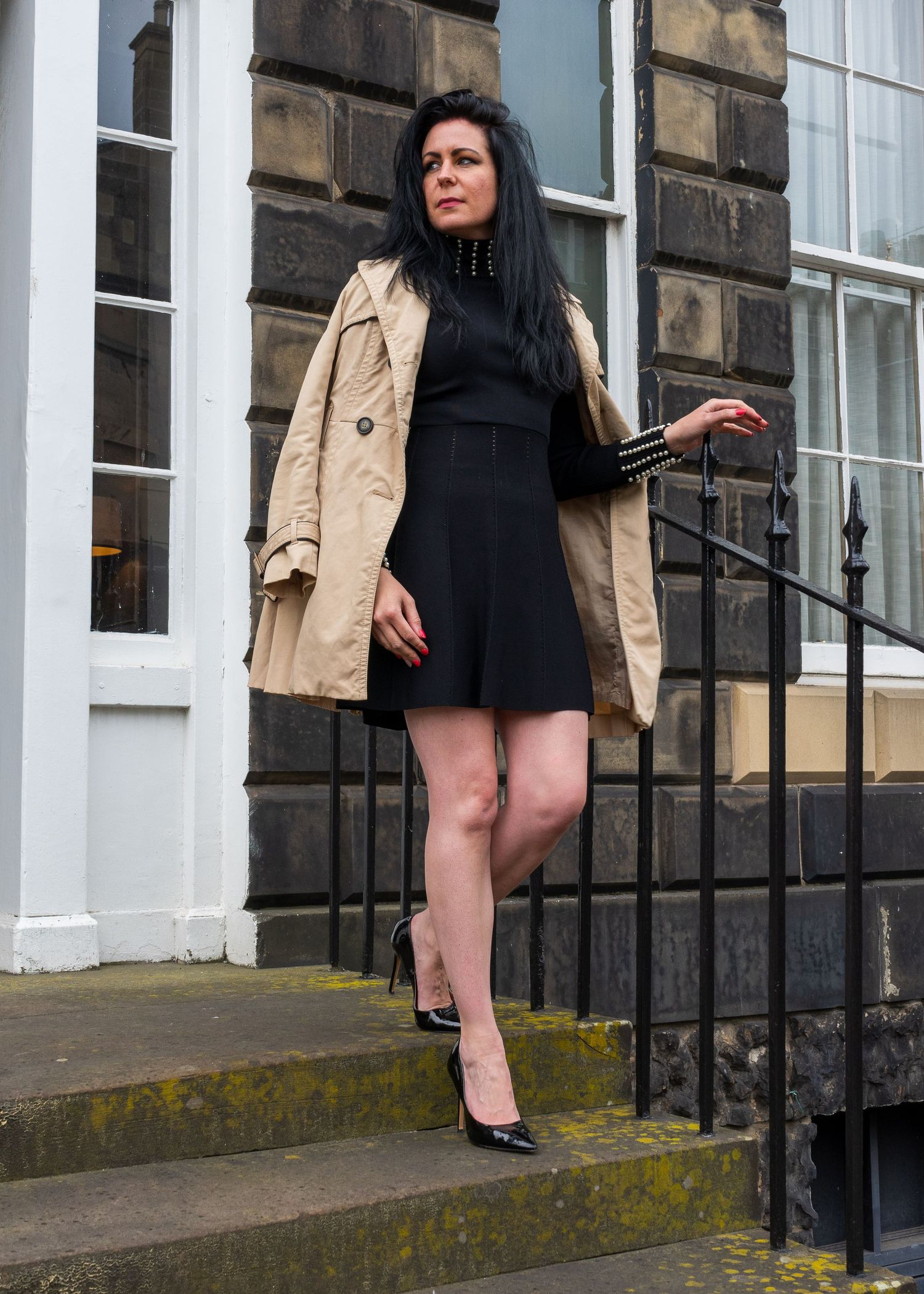 Person in black outfit and beige coat poses on stone steps of historic building.