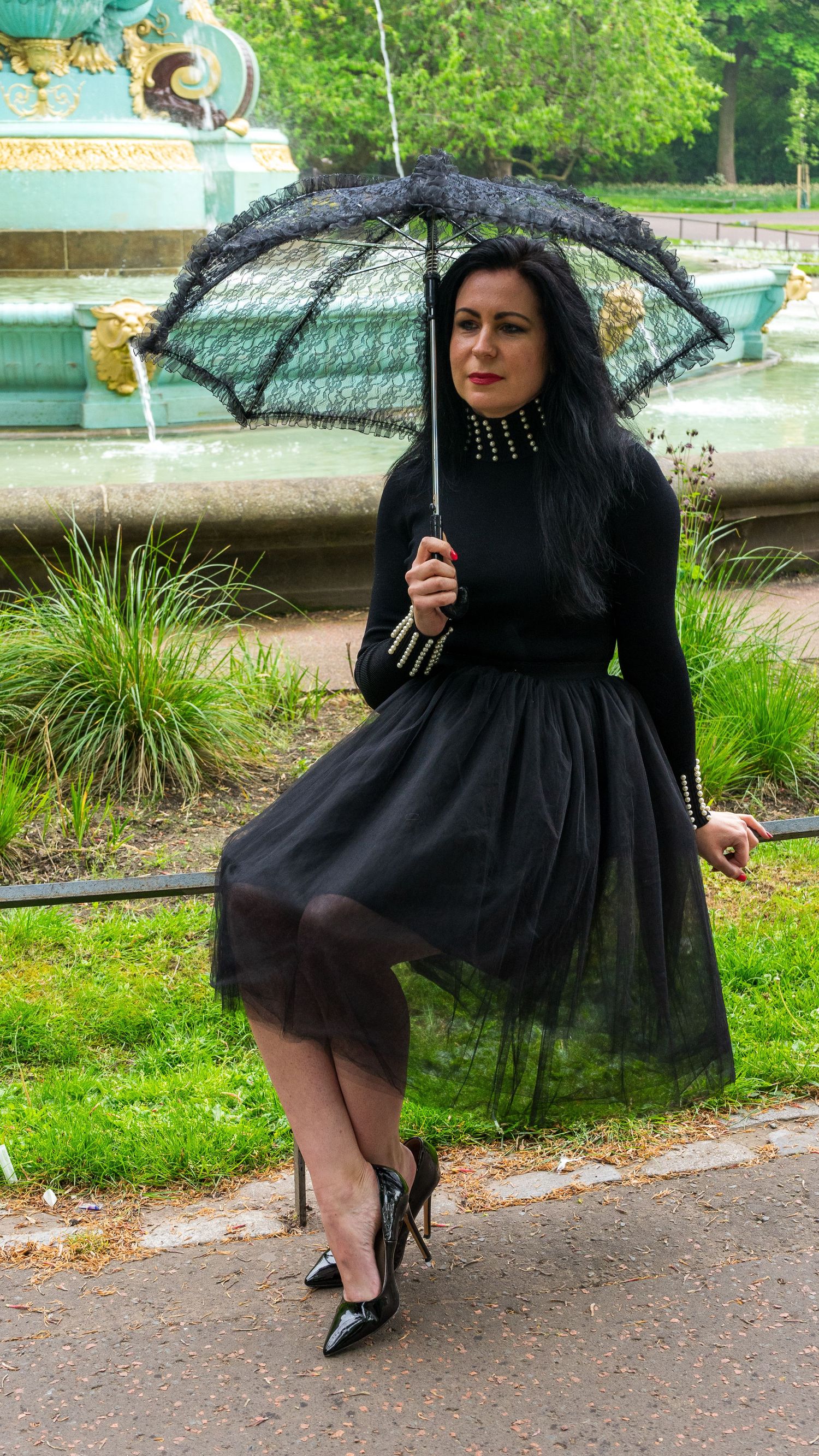 Figure in flowing black dress twirls with lace umbrella in garden setting.