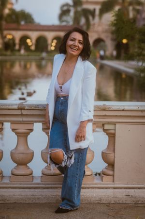 Casual chic look with white blazer and distressed jeans posing by ornate balustrade at sunset.