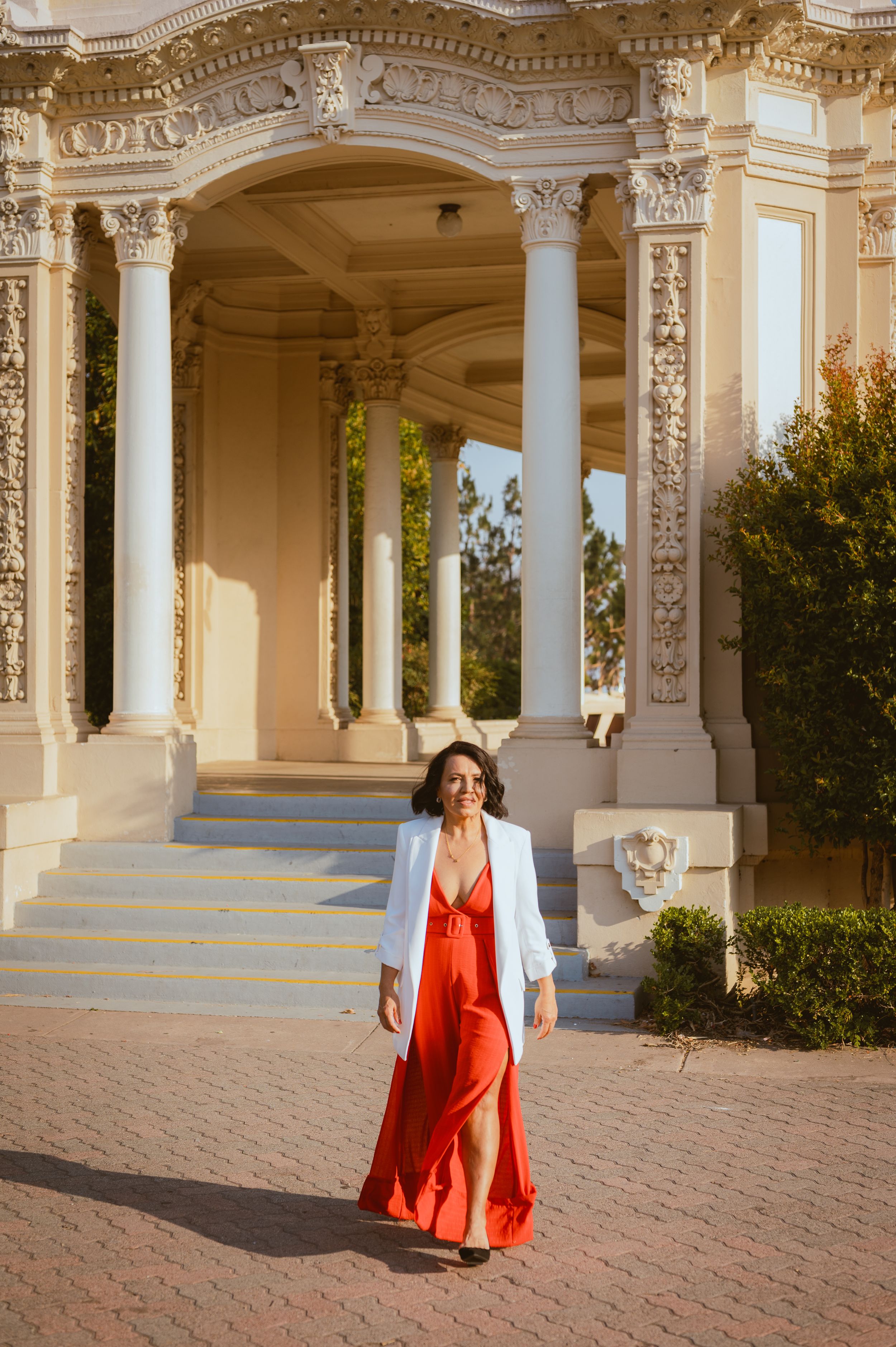 Woman in flowing red dress and white blazer poses elegantly at ornate columned building entrance.