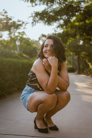 Woman in casual summer outfit crouches playfully on garden path during sunset photoshoot.