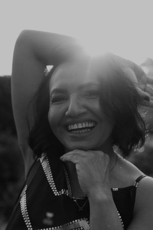 A joyful portrait in black and white showing a radiant smile and beaded necklace against a dark background.