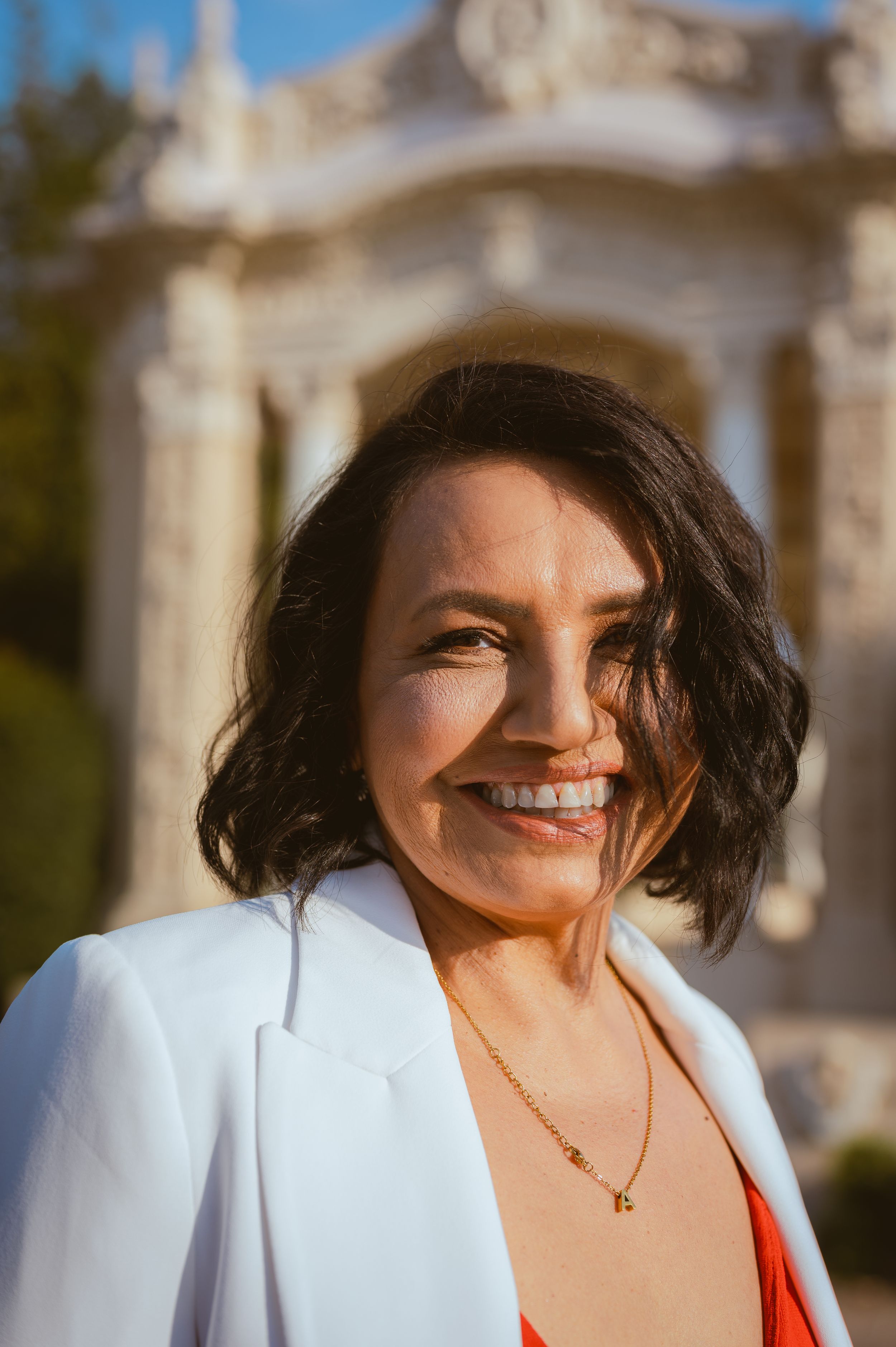 Portrait of woman in white blazer and red dress smiling in front of classical architecture.