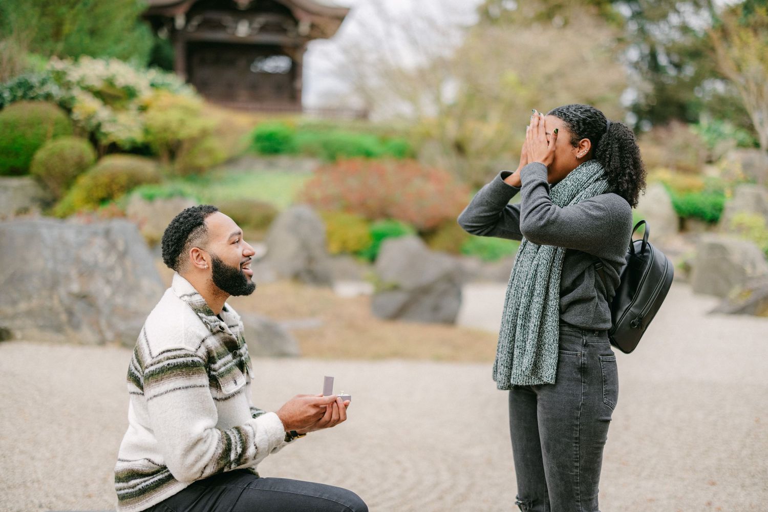A surprise proposal moment captured at a Japanese garden with one person kneeling and another covering their mouth in joy.