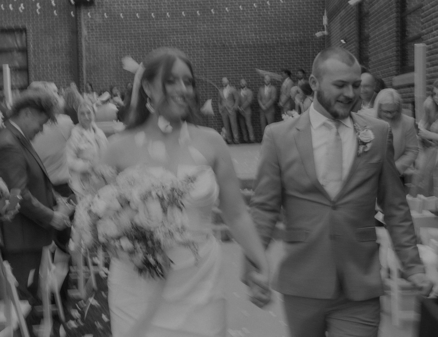 A bride and groom embrace with coral dahlia flowers in the foreground of a moody wedding photo.