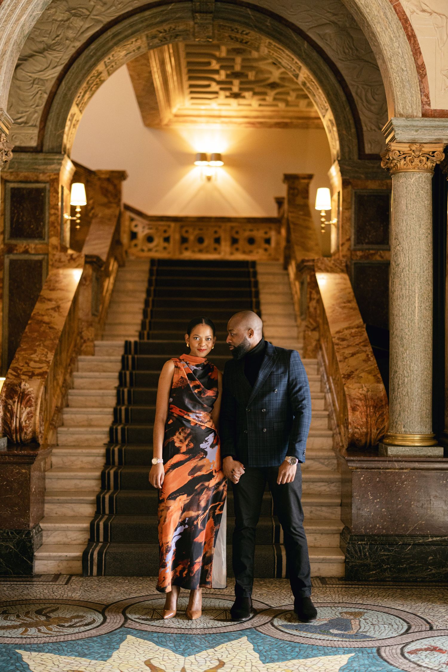 A couple poses elegantly beneath an ornate archway and grand staircase in a historic building.