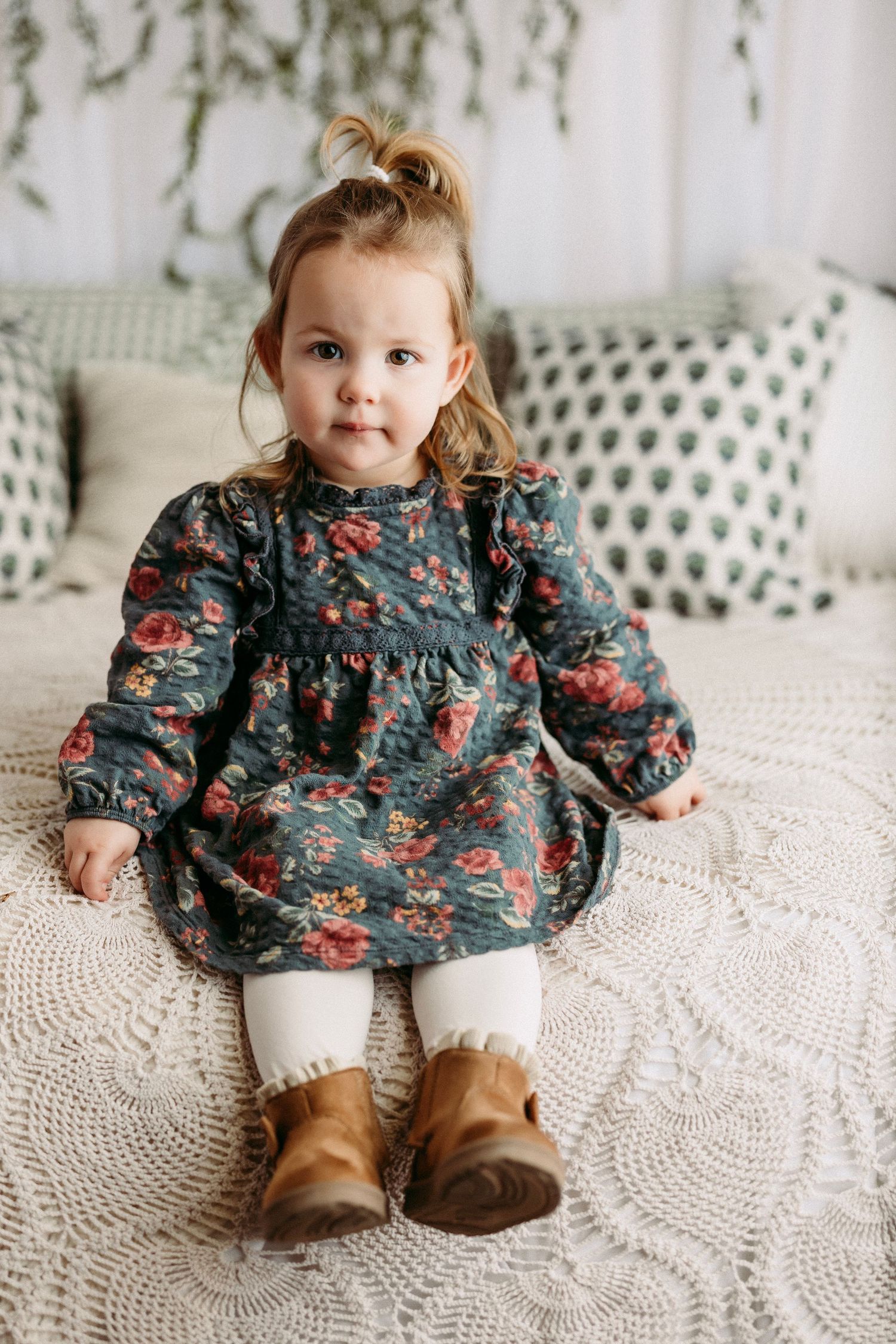 A small child in a floral dress and boots sits on a white crocheted blanket in a series of candid poses.