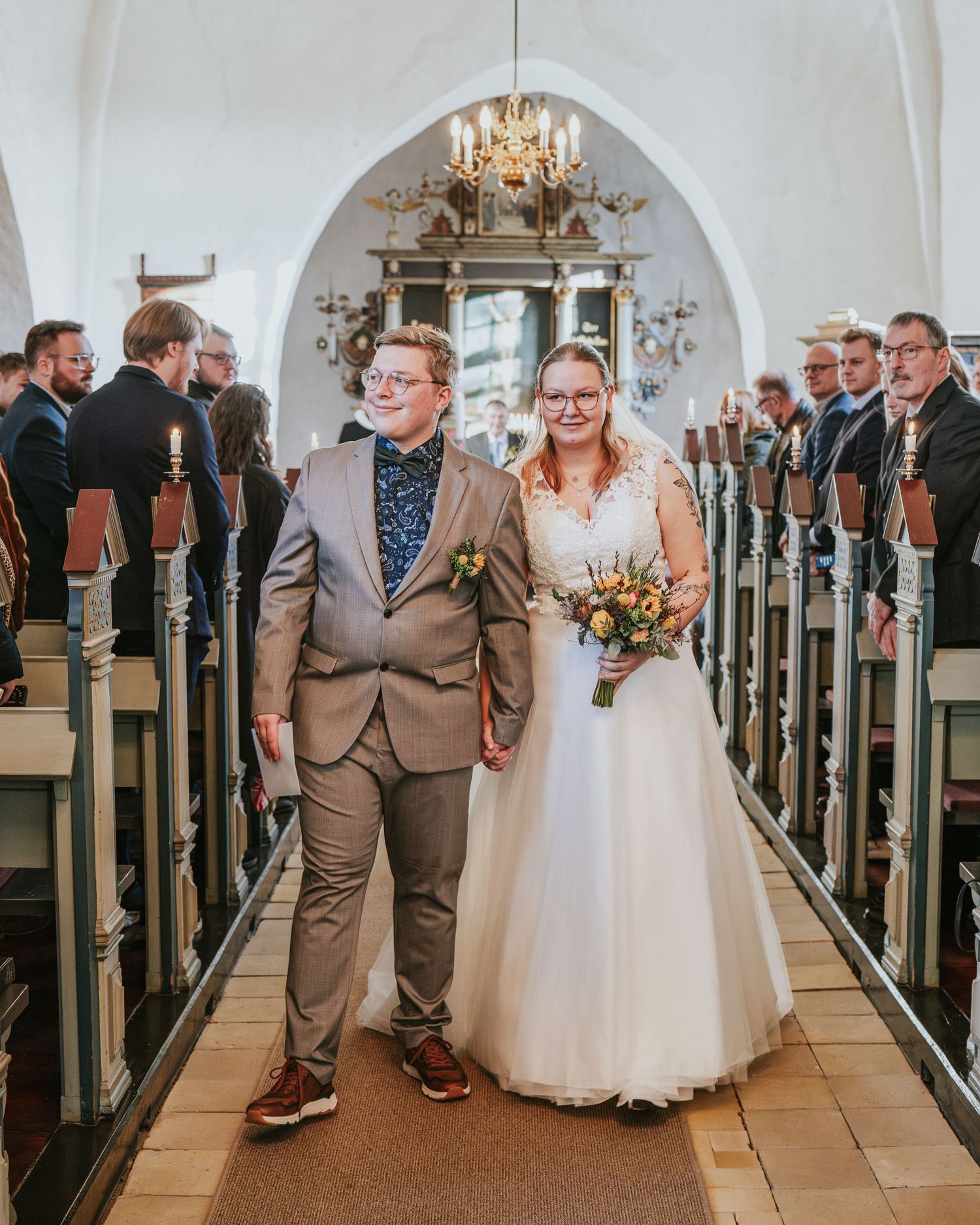 Wedding ceremony in a traditional church with wooden pews and chandelier lighting.