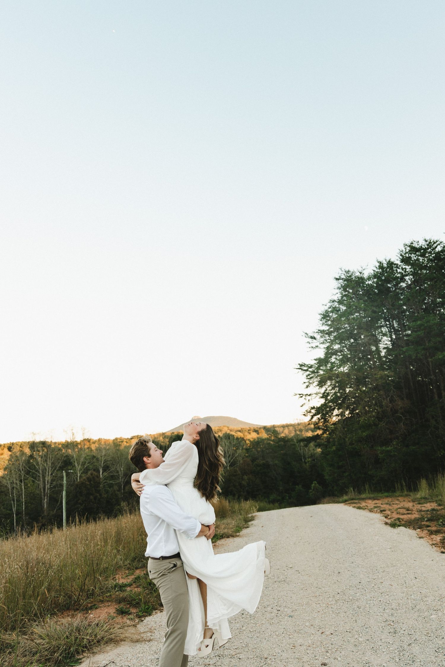 Romantic embrace on country road with flowing white dress against natural landscape at sunset.
