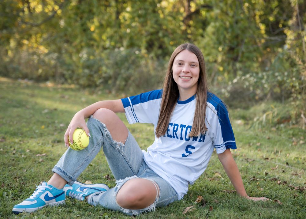 Softball player in a white and blue uniform sitting on grass with a yellow softball.