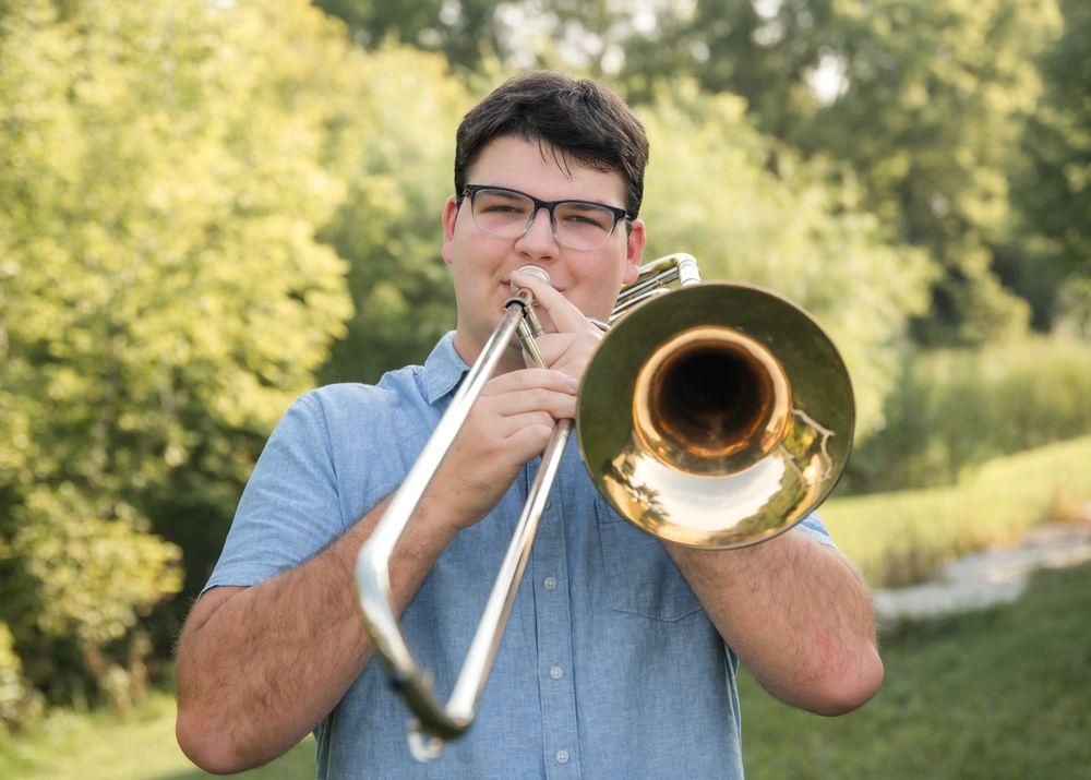 Musician plays brass trombone outdoors in casual blue shirt against green foliage background.