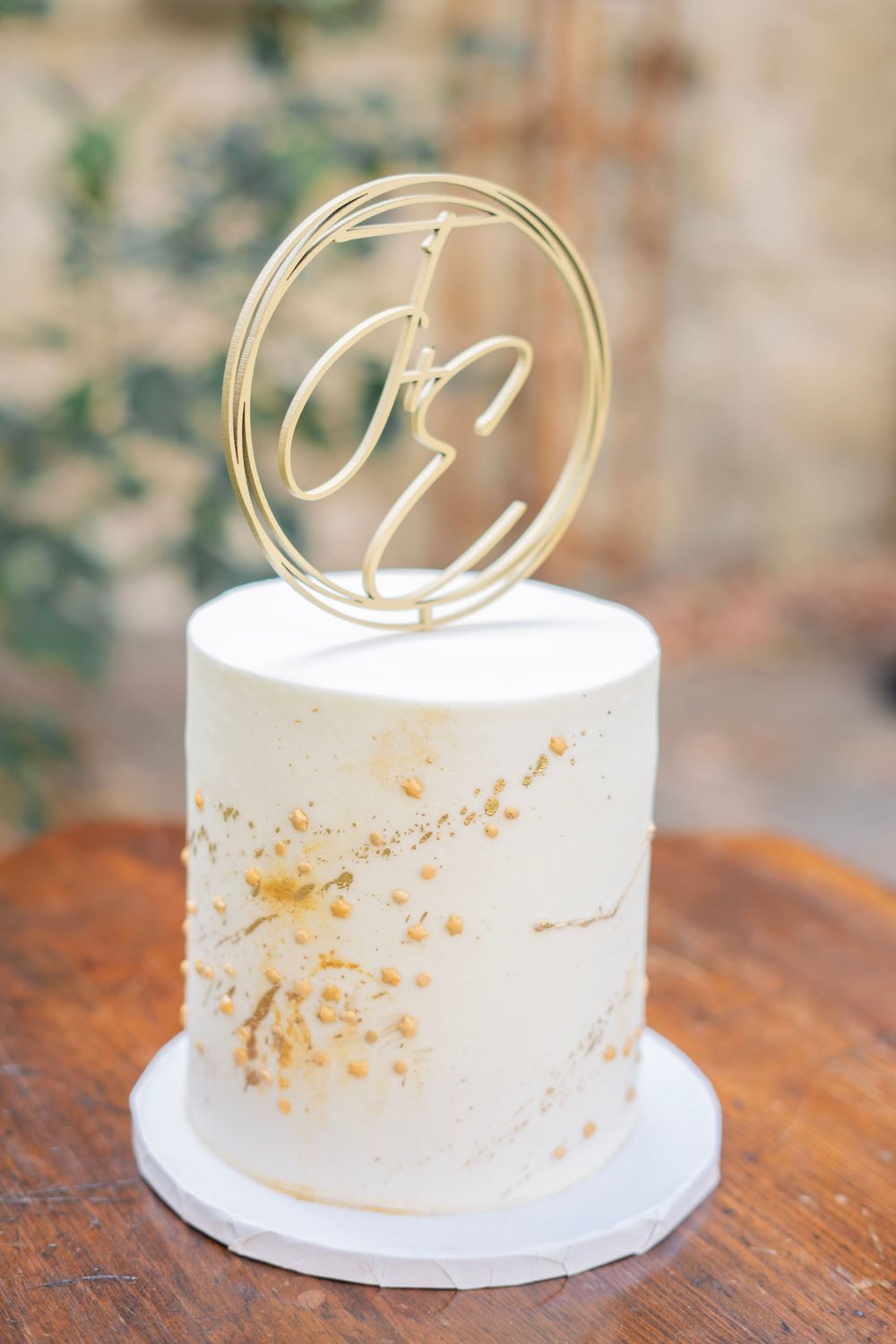 Minimalist white single-tier wedding cake with gold monogram letter E topper and gold splatter details.
