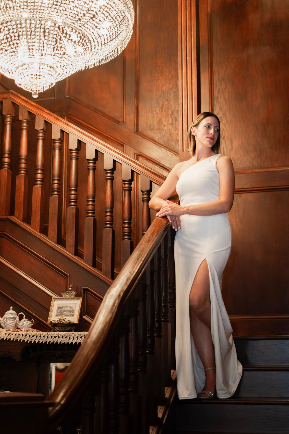 Bride in sleek white one-shoulder gown with thigh slit poses on ornate wooden staircase under crystal chandelier.