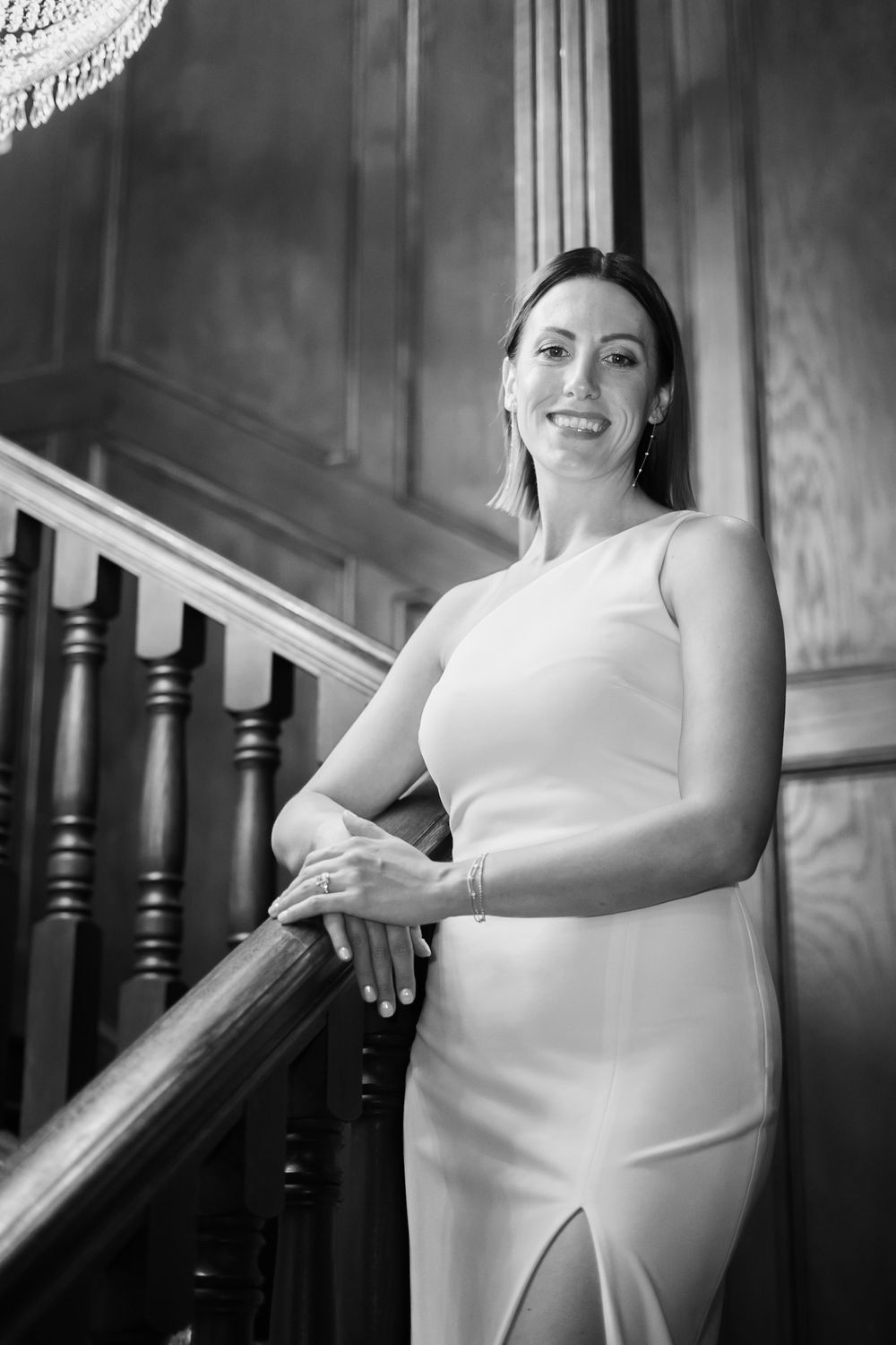 Elegant bride in white one-shoulder gown posing on classic wooden staircase in black and white photo.