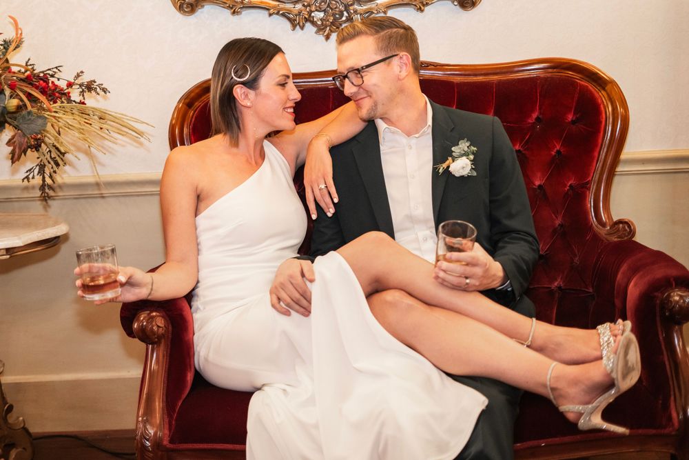 Bride and groom sitting on ornate red velvet sofa, smiling at each other while holding drinks.