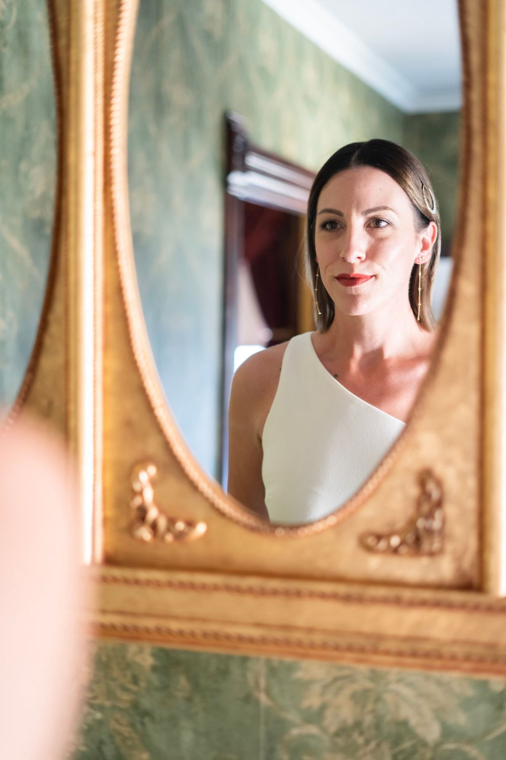 Elegant bride with red lips in white one-shoulder dress gazing away in ornate gold oval mirror reflection.