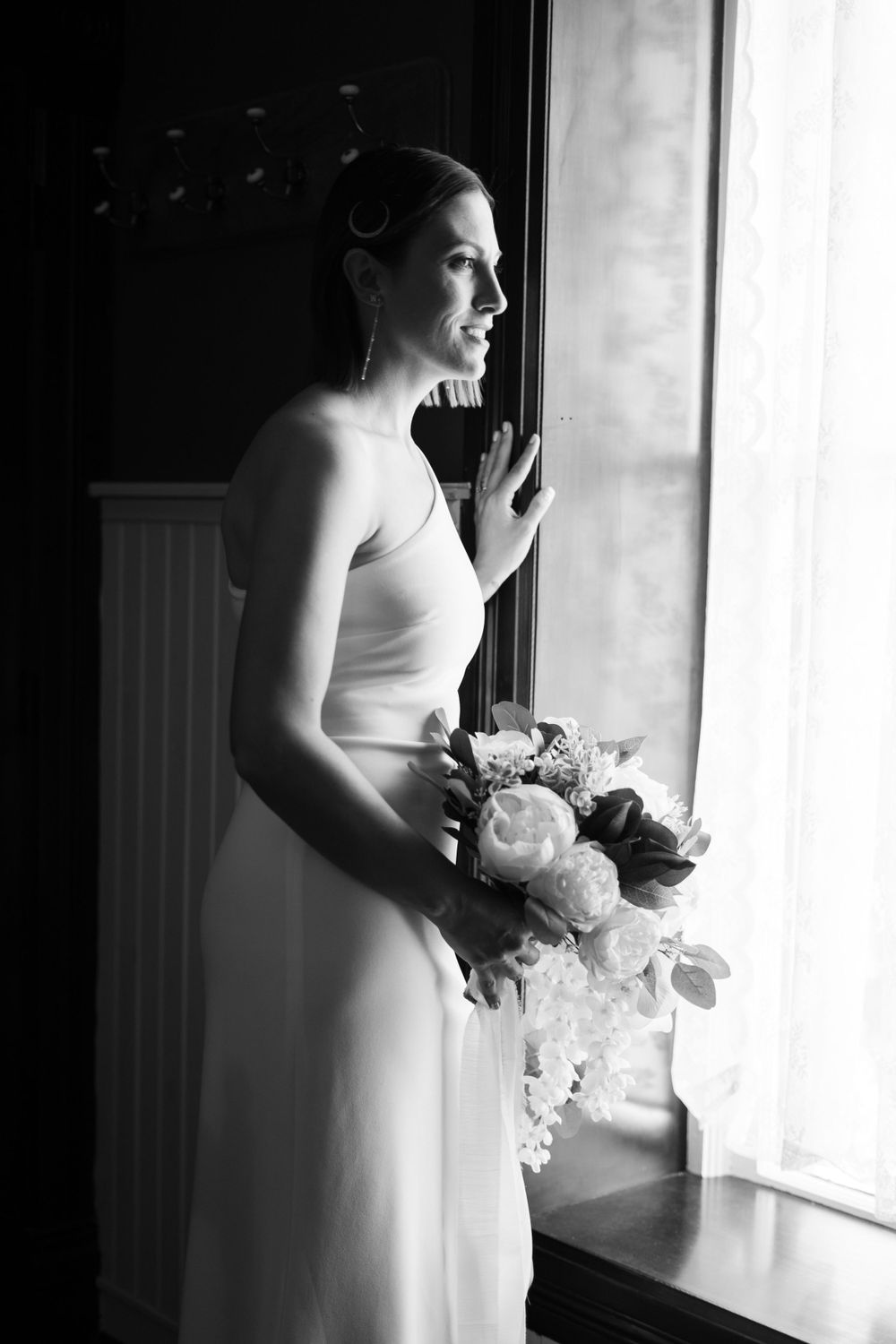 Elegant bride in strapless white gown holding floral bouquet gazing out window in black and white wedding photo.