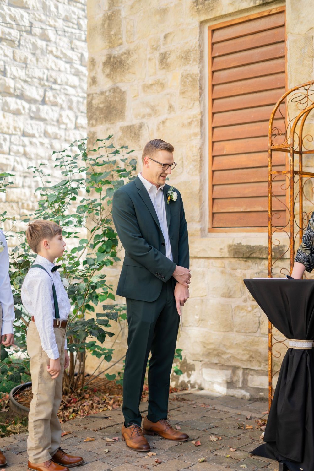 Groom in dark green suit stands at altar with young boy in suspenders at outdoor ceremony.