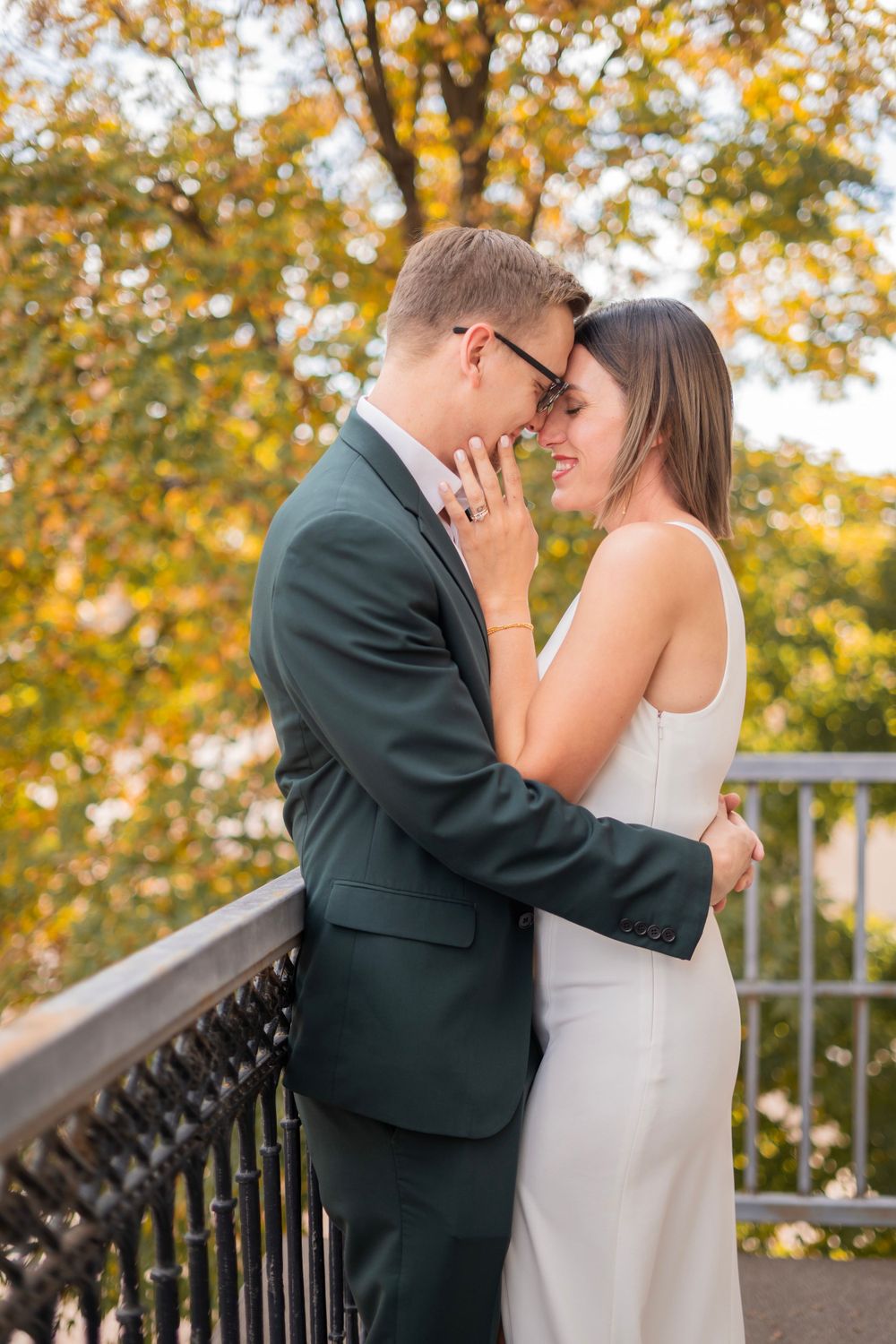 Bride and groom share an intimate nose-to-nose moment on a balcony with fall foliage backdrop.